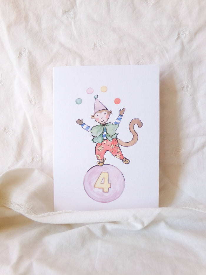 Card with a monkey character on a pink ball with the number 4, on a beige fabric background.