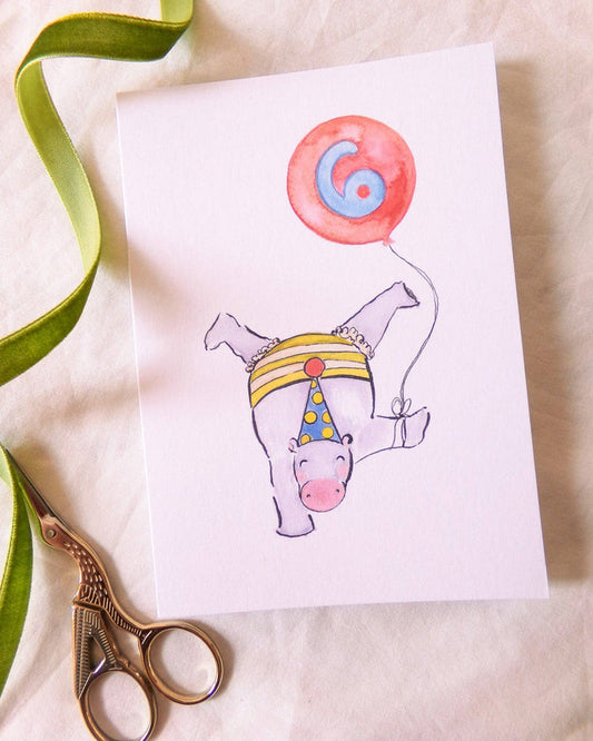 Illustration of a hippo with a party hat and balloon on a card, with scissors and ribbon on a beige background.