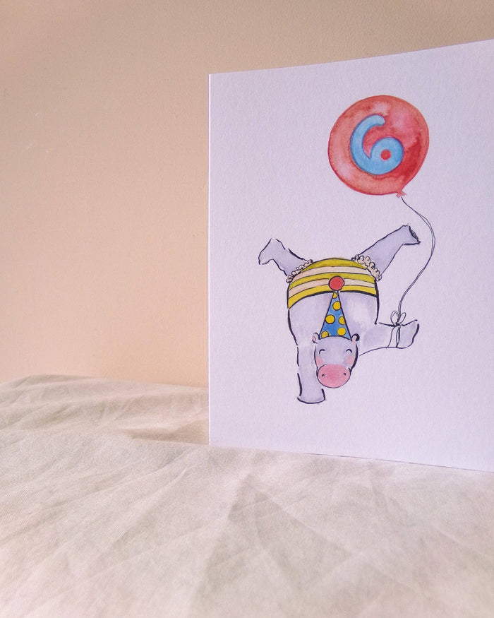 A white greeting card standing upright on a light fabric surface against a warm beige background. The card features a whimsical watercolor illustration of a white pig wearing a yellow and blue party hat and holding a red balloon with a blue spiral design.