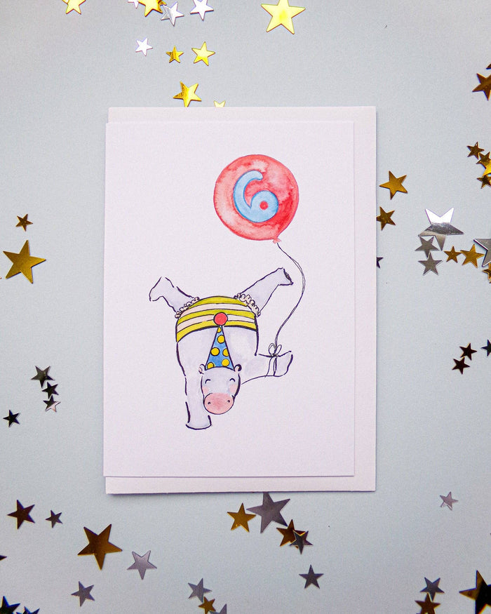 A white greeting card featuring a whimsical illustration of a white horse upside down holding a red balloon with a blue number 6, displayed on a light gray background decorated with scattered gold and silver star confetti.