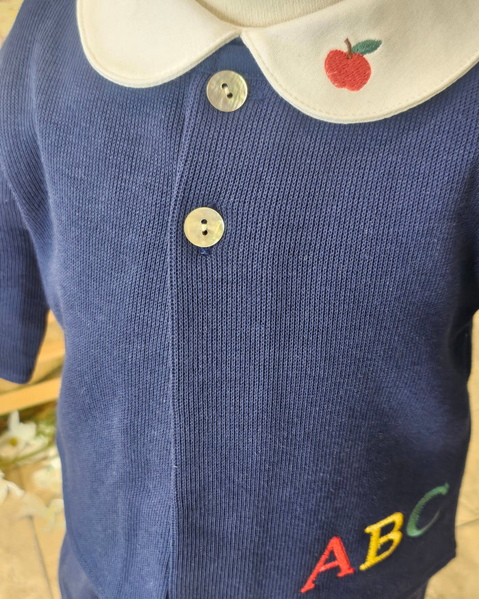 Close-up of a navy blue ribbed knit baby top with a white collar featuring a red apple embroidery, two silver buttons, and colorful embroidered letters 'ABC' on the chest.