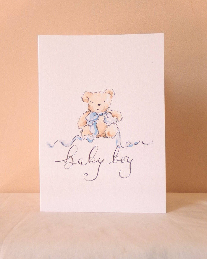 A vertical baby shower card featuring a watercolor illustration of a teddy bear wearing a blue bow, above the cursive text 'baby boy' in dark blue ink, set against a warm beige background.
