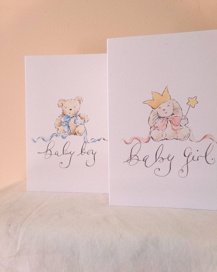 Two white baby shower cards standing upright on a white fabric surface against a warm beige wall. The left card says 'baby boy' with a teddy bear illustration in blue.