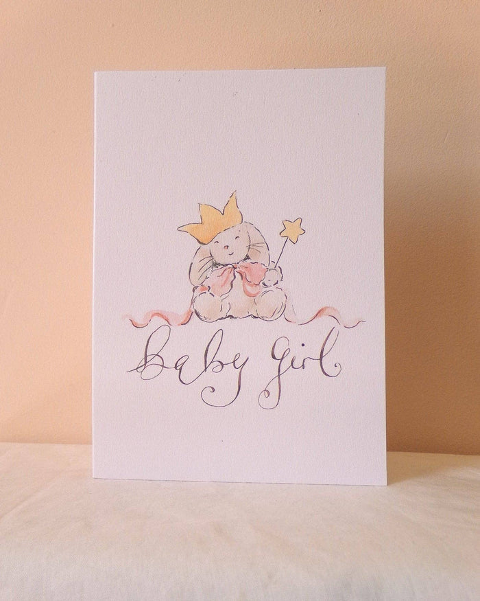 A white baby shower card featuring a watercolor illustration of a teddy bear wearing a gold crown and holding a pink bow, with the words 'baby girl' written in elegant cursive script below, set against a warm beige background.