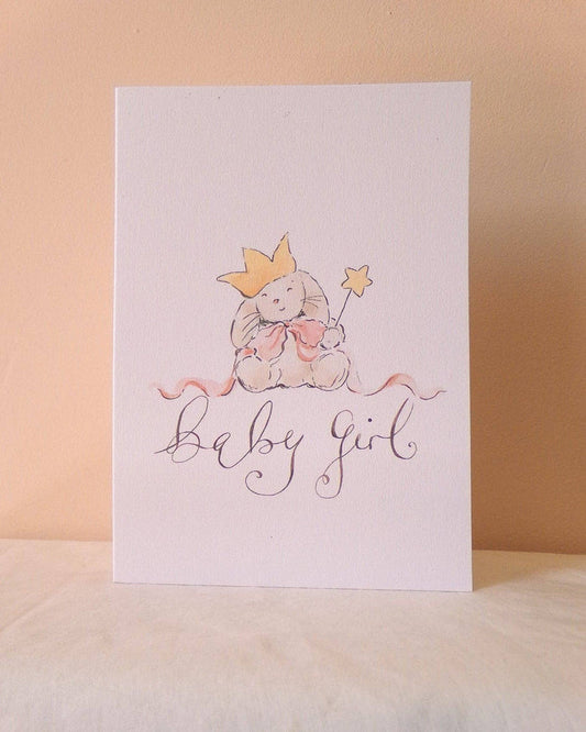 A white baby shower card featuring a watercolor illustration of a teddy bear wearing a gold crown and holding a pink bow, with the words 'baby girl' written in elegant cursive script below, set against a warm beige background.