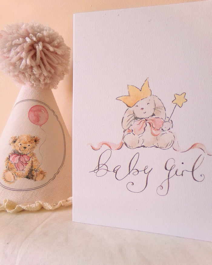 A baby girl birthday card featuring a watercolor illustration of a bunny wearing a gold crown and holding a star wand, with the words 'baby girl' written in elegant cursive. A matching baby hat with a pom-pom is partially visible on the left.