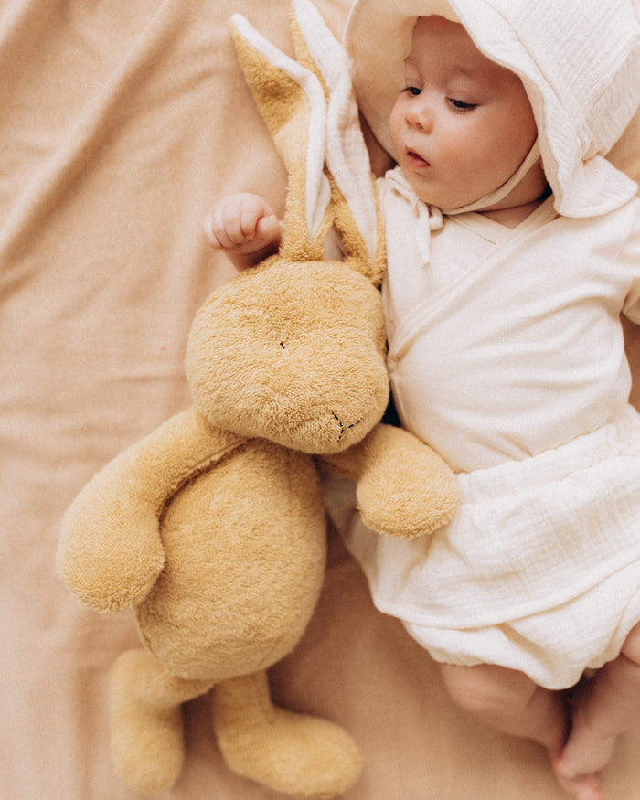 Baby in a white hooded outfit holding a plush bunny toy on a beige blanket