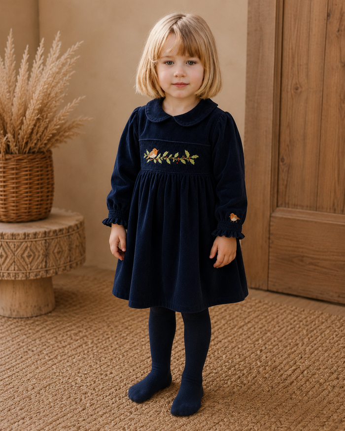 Young girl in a navy dress with floral embroidery standing in a room with a wooden door and woven basket.