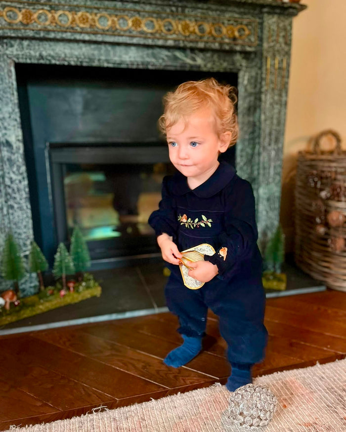A toddler with curly blonde hair wearing a navy blue embroidered jumpsuit and blue socks stands in front of a fireplace, holding a small yellow-green toy.