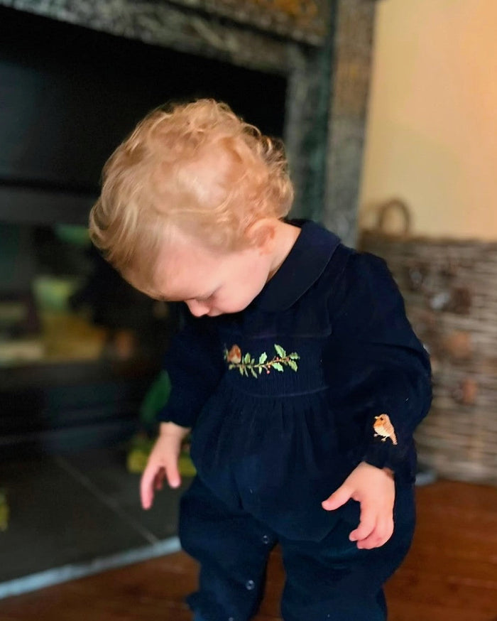A toddler with curly blonde hair wearing a navy blue velvet jumpsuit with green and orange floral embroidery on the chest, standing in a home interior near a fireplace.