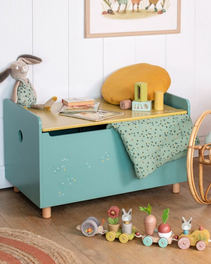 Children's toy storage bench with a wooden top and floral design on the sides, surrounded by toys in a room.