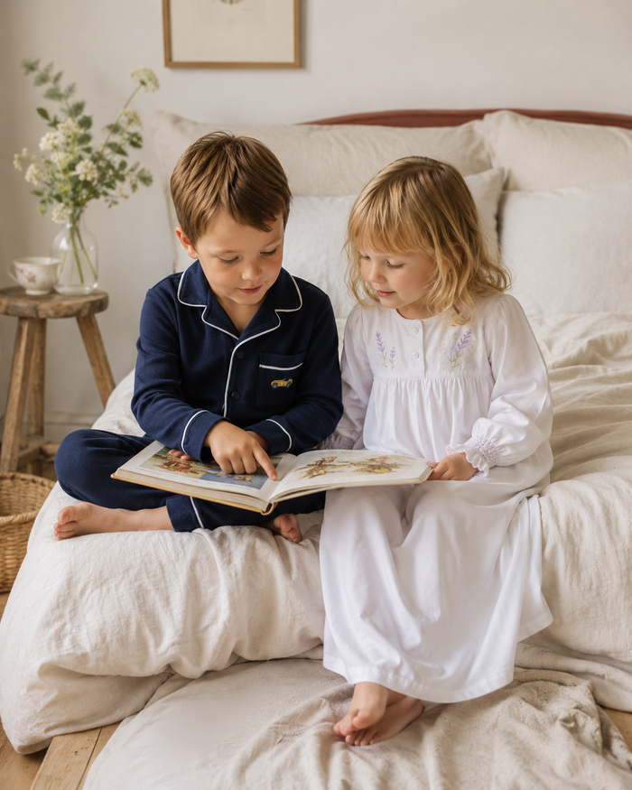 Two children sitting on a bed reading a book in a cozy bedroom setting. The boy wears a navy blue pajama set with white piping, and the girl wears a long white nightgown.