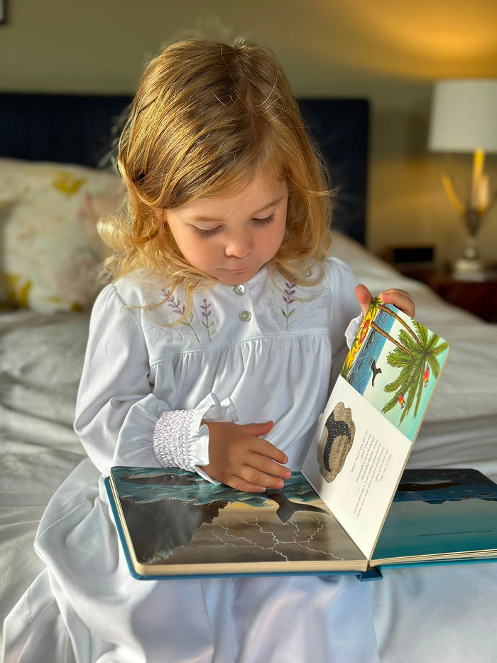 A young girl with blonde hair sits on a bed, reading an open illustrated picture book. She is wearing a white long-sleeve nightgown with floral embroidery on the chest.
