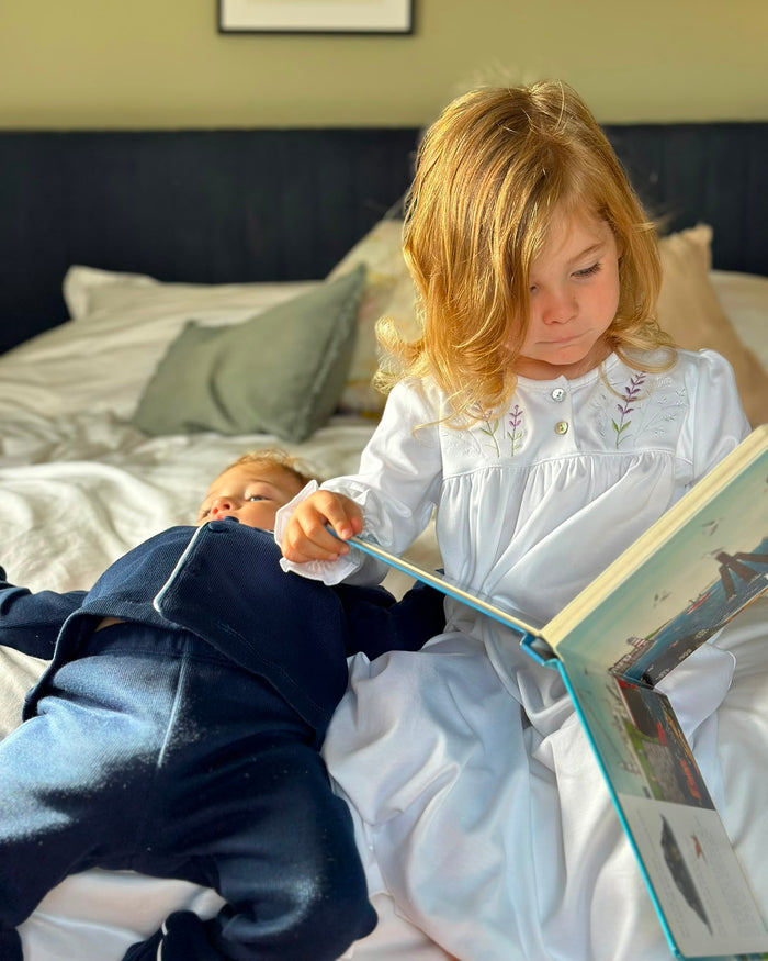 A young girl in a white embroidered nightgown sits on a bed reading a book, while a baby in a navy blue outfit lies beside her on the bedding.