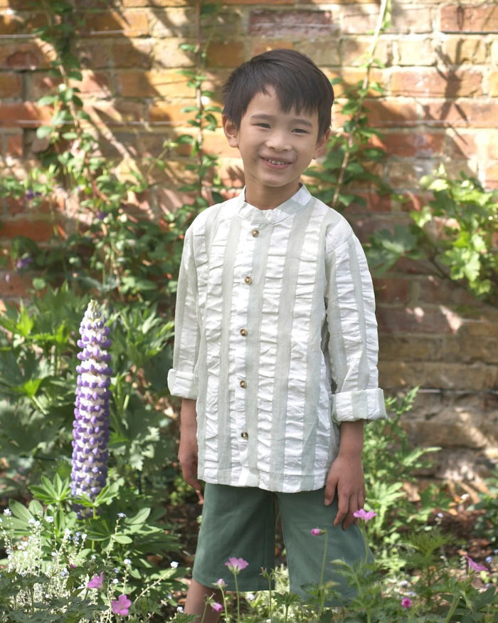 Child wearing a white textured shirt and green shorts standing in a garden with flowers and a brick wall.