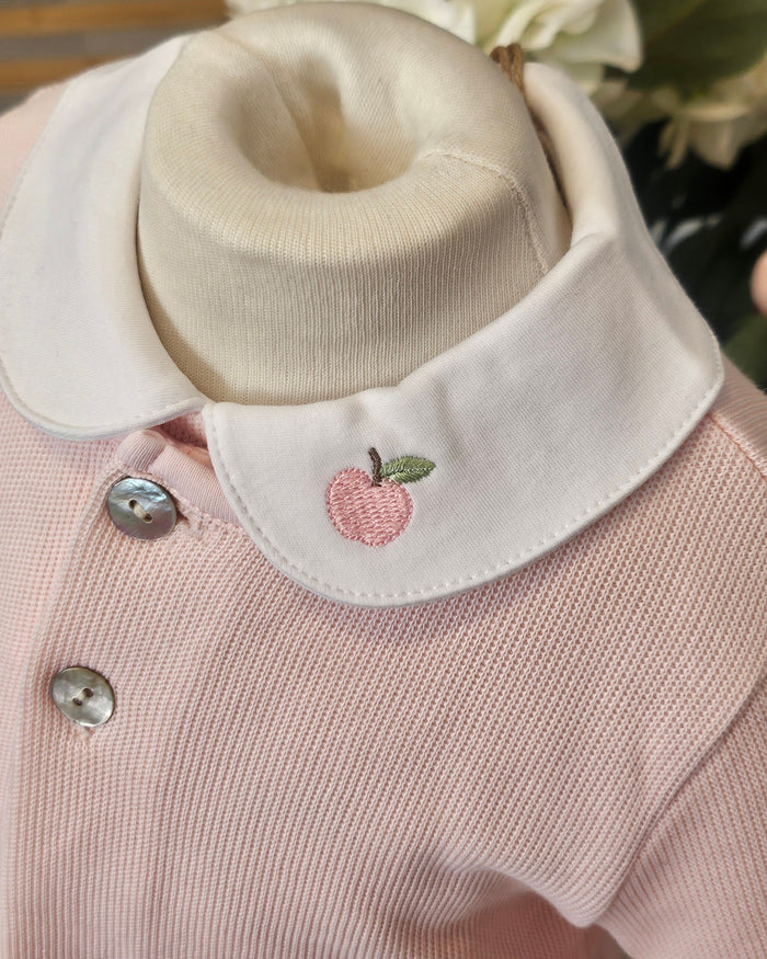 A close-up of a baby clothing display showing a light pink ribbed knit top with a wide cream-colored collar. The collar is embroidered with a small pink apple and green leaf, and has silver buttons.