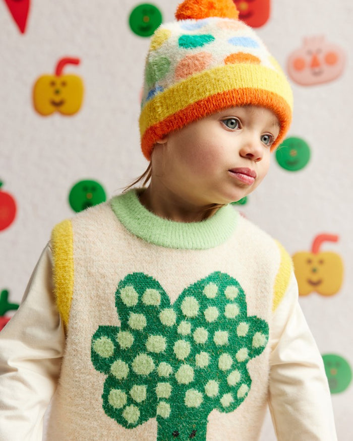 Child wearing a colorful knit hat and sweater with a green broom design against a patterned background.
