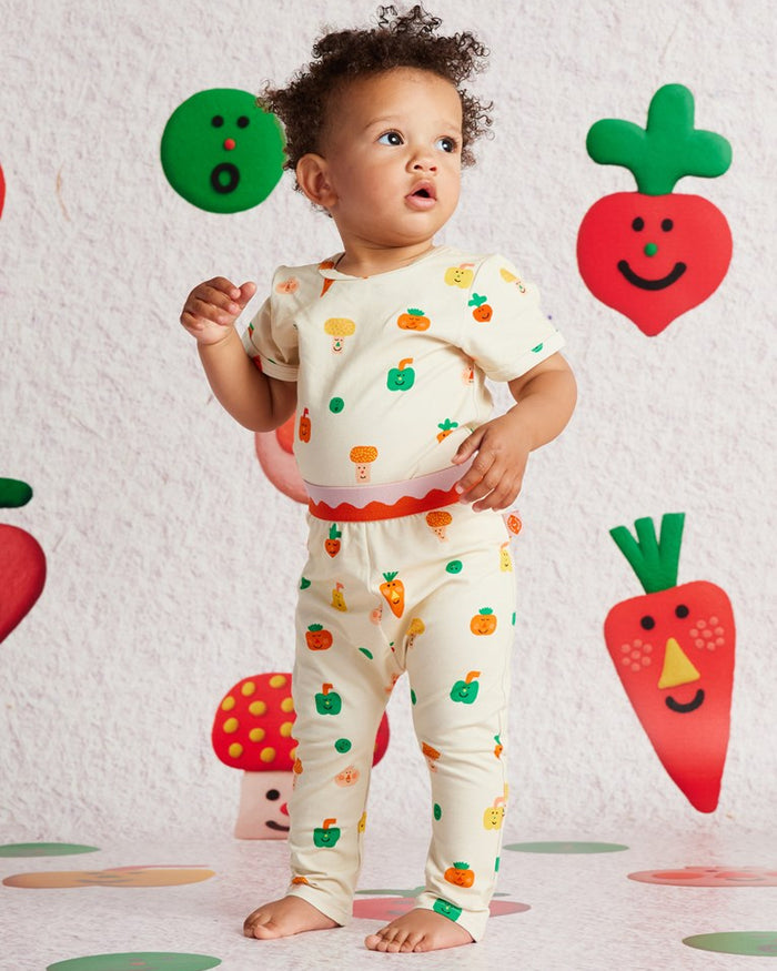 Baby wearing a colorful outfit with vegetable patterns against a wall with vegetable-themed decorations.