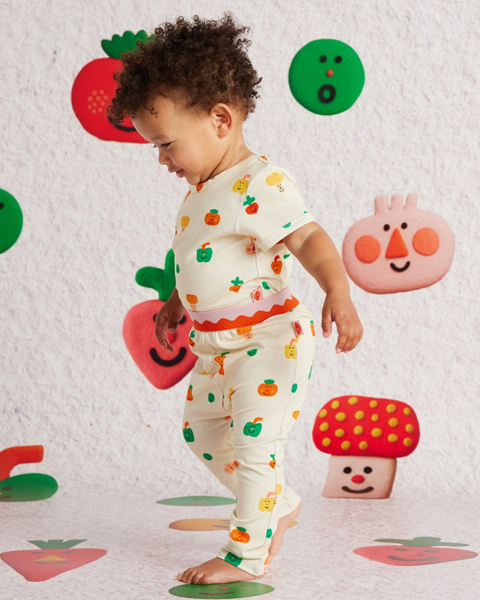 Child in a colorful outfit standing on a patterned floor with cartoonish vegetables and fruits.