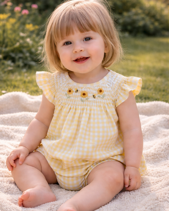 Child wearing a yellow checkered outfit with sunflower details, sitting on a blanket outdoors.