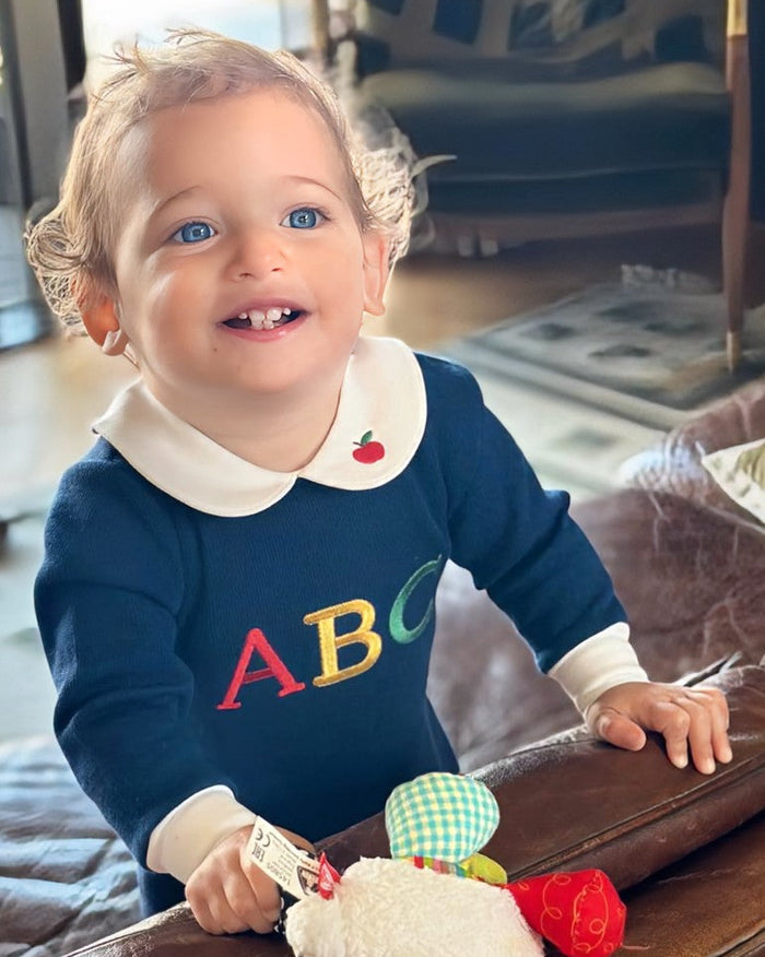 A smiling toddler standing indoors, wearing a navy long-sleeve top with a cream Peter Pan collar, white cuffs, and colorful letters 'ABC' across the chest. The child is holding a plush toy in front of a brown leather surface, with a blurred living room background.