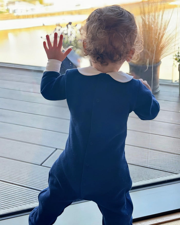 A toddler seen from behind wearing a navy blue footless one-piece jumpsuit with a contrasting white Peter Pan collar, standing on a wooden deck near a window.