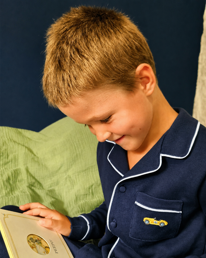 A young boy with short blond hair wearing navy blue pajamas with white piping and a small yellow car embroidery on the chest pocket, looking down at a book in his hands.
