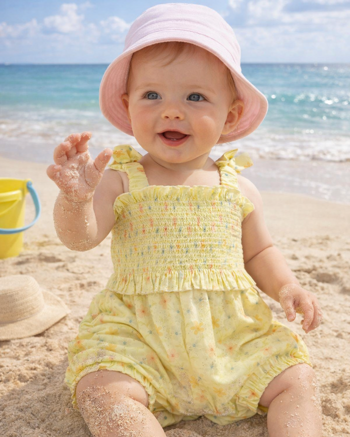 Baby in a yellow outfit and pink hat sitting on a sandy beach with ocean in the background