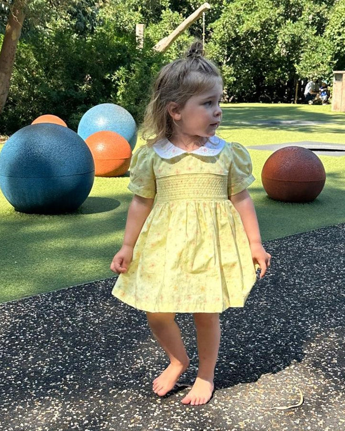A toddler girl wearing a light yellow smocked dress with a white Peter Pan collar stands barefoot on an outdoor playground path. Large colorful exercise balls and green grass are visible in the background.