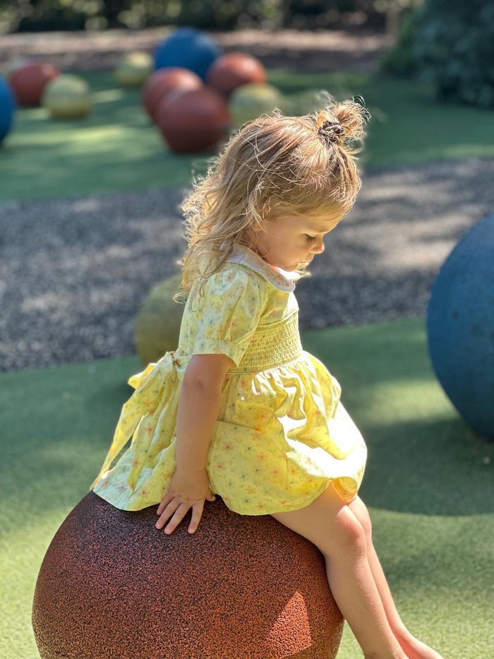 A young girl with blonde hair in a small bun sits on a large brown ball in an outdoor playground, wearing a yellow short-sleeve dress with a white pattern.