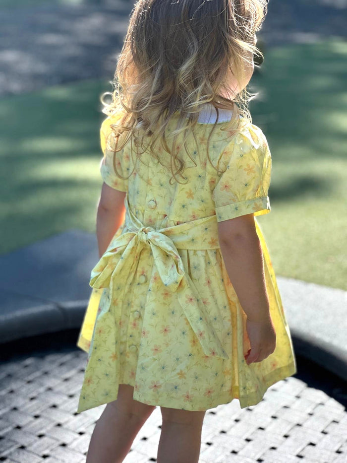 A young girl viewed from behind wearing a yellow short-sleeve dress with a small floral print and a tie waist, standing outdoors on a paved surface with grass in the background.