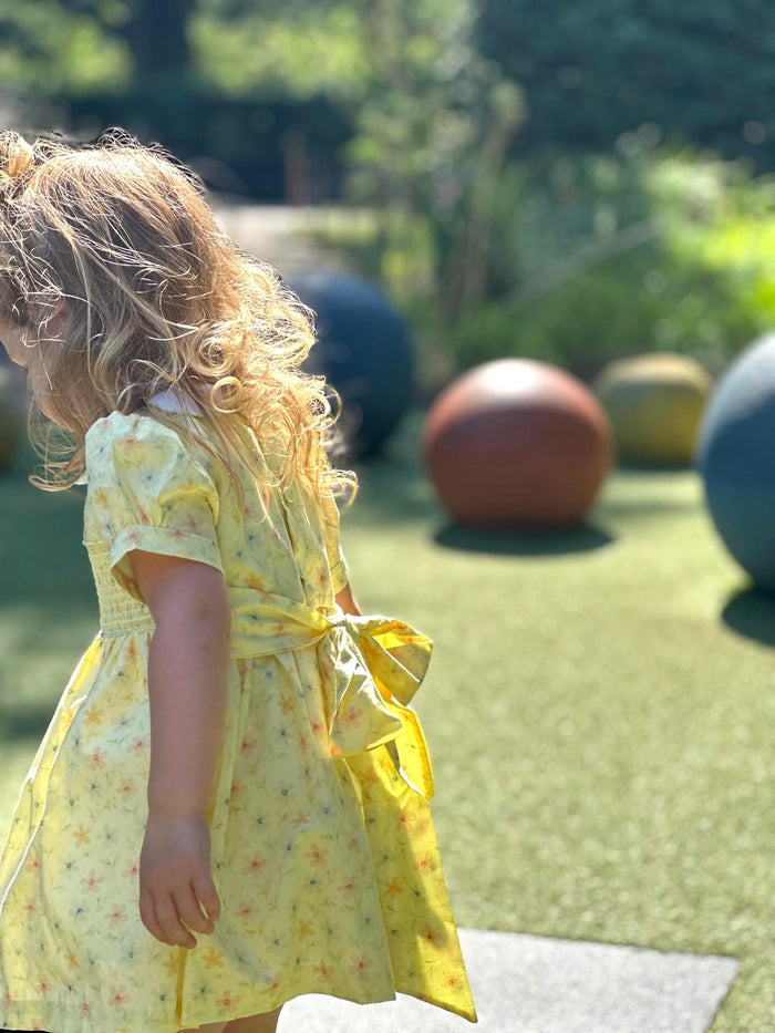 A young child with curly blonde hair seen from the side, wearing a yellow short-sleeve dress with a small floral print and a tied waist bow, standing on artificial grass in an outdoor playground with colorful exercise balls in the background.
