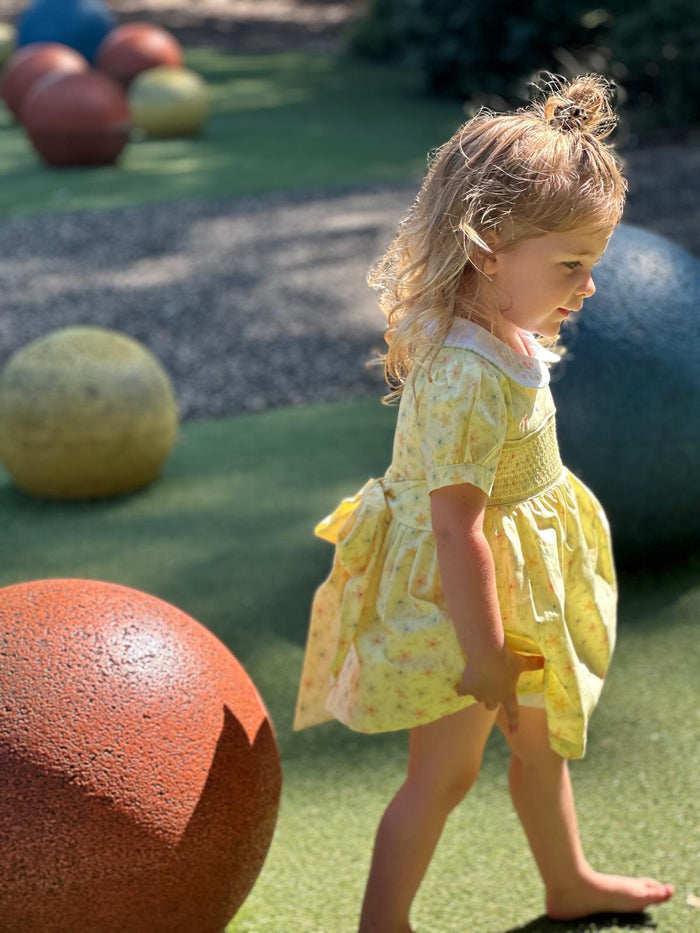 A young toddler girl with blonde hair in a small bun wearing a yellow patterned dress, walking barefoot on green artificial turf in an outdoor playground setting with large decorative spheres.