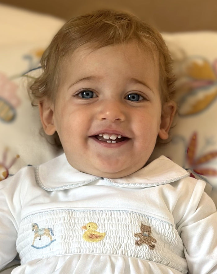A close-up of a smiling toddler wearing a white long-sleeve dress with a Peter Pan collar and smocked details featuring embroidered toys.