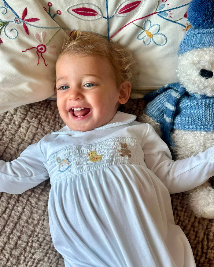 A baby girl lying on her back on a textured brown blanket, wearing a white long-sleeve baby smocked dress with a smocked chest panel. She is smiling broadly.