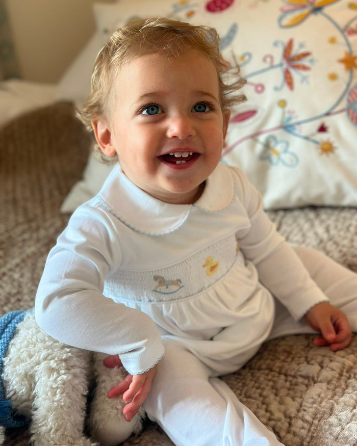 A smiling toddler with blonde hair and blue eyes wearing a white long-sleeve baby smocked jumpsuit, sitting on a textured beige blanket with a floral pillow in the background.