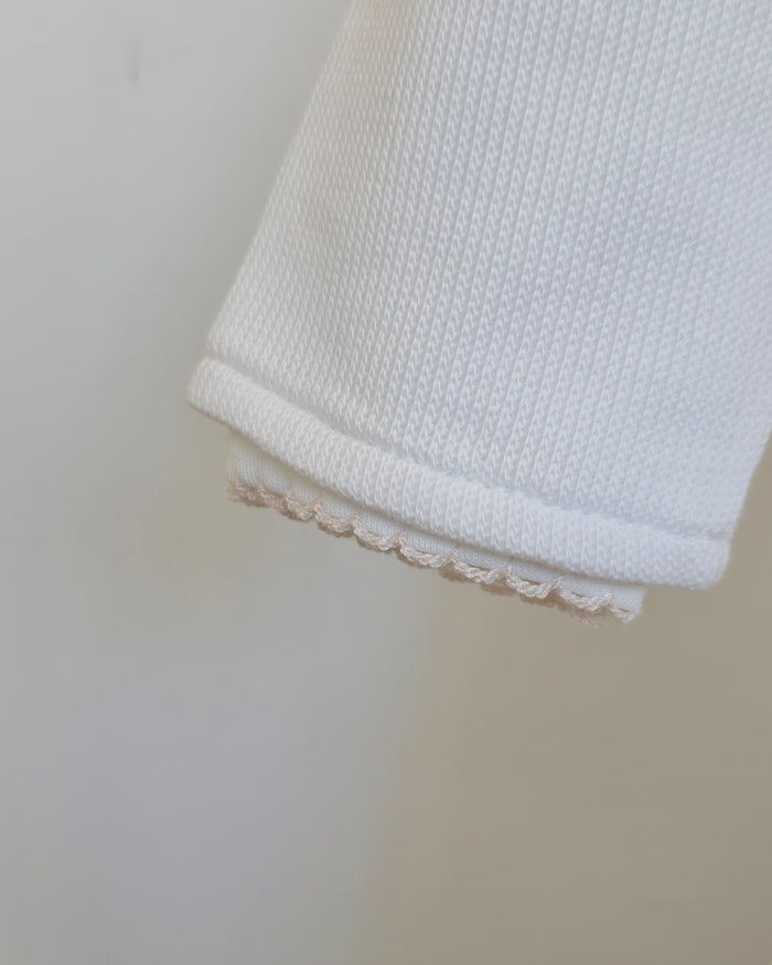 Close-up of a white ribbed knit fabric cuff with a delicate beige scalloped trim along the edge, set against a plain light beige background.