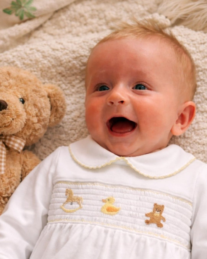 Baby in a white outfit with embroidered designs, lying on a soft surface next to a teddy bear.