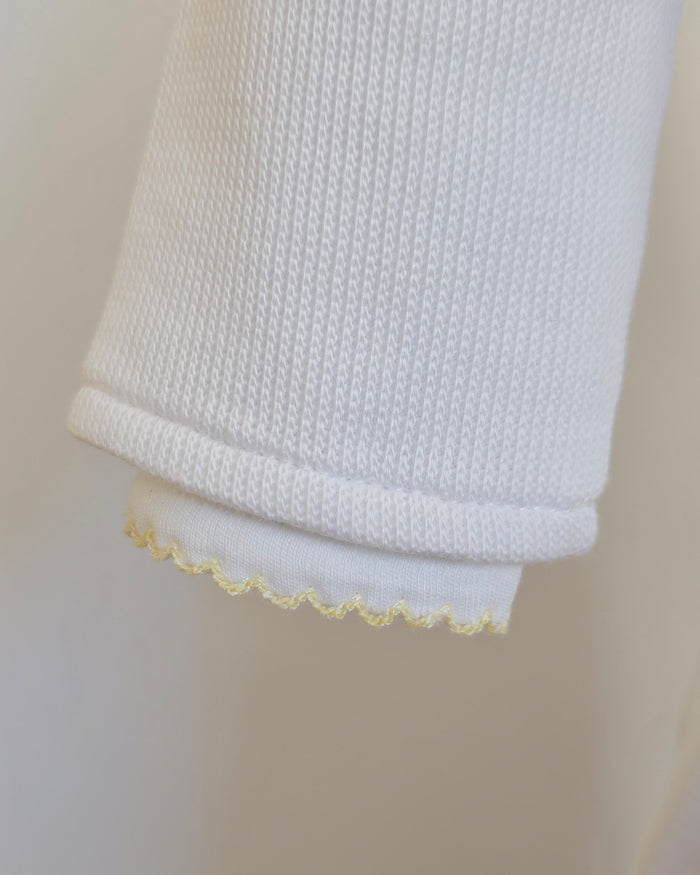 Close-up of a white ribbed knit sleeve or cuff with a delicate yellow scalloped trim along the inner edge, shown against a soft light beige background.