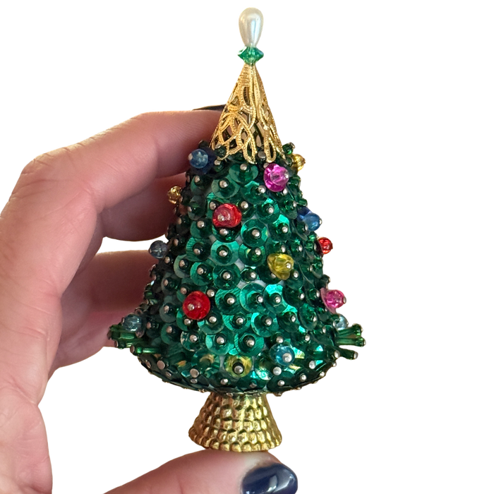 Hand-held close-up of a vintage green sequin Christmas tree ornament showing colorful pinned beads, an openwork gold-tone top, and a textured base.