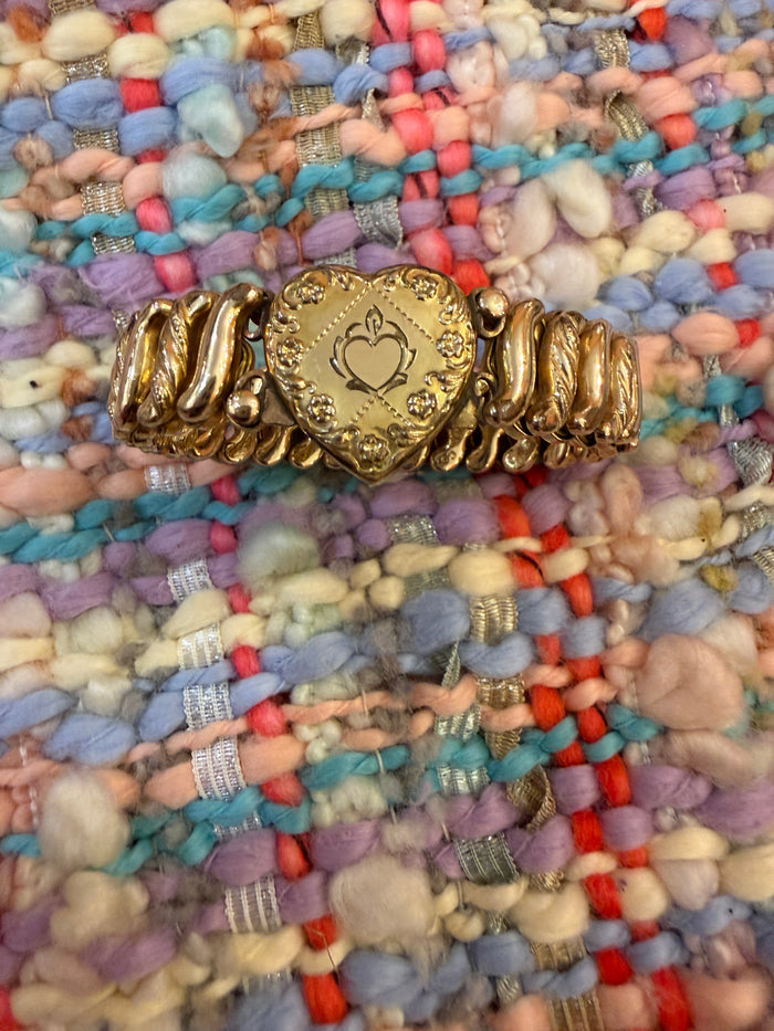 Vintage gold-tone expansion bracelet with ornate embossed heart clasp displayed against a colorful woven textile.