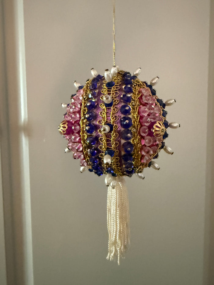 Close-up of a vintage purple and pink beaded ornament showing gold-tone floral details, decorative trim, pearl-look beads, and an ivory tassel.