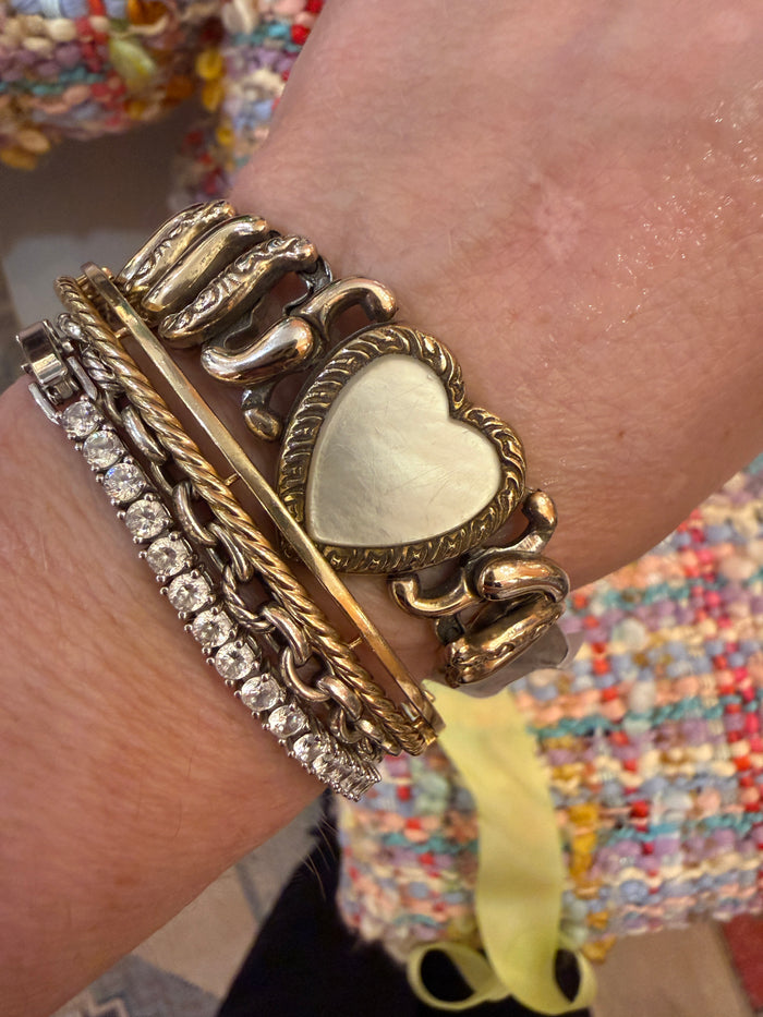 A close-up of a person's wrist wearing a stack of chunky vintage-style bracelets, including a heart-shaped charm bracelet, a rope chain bracelet, and a silver-toned chain bracelet with clear stones.