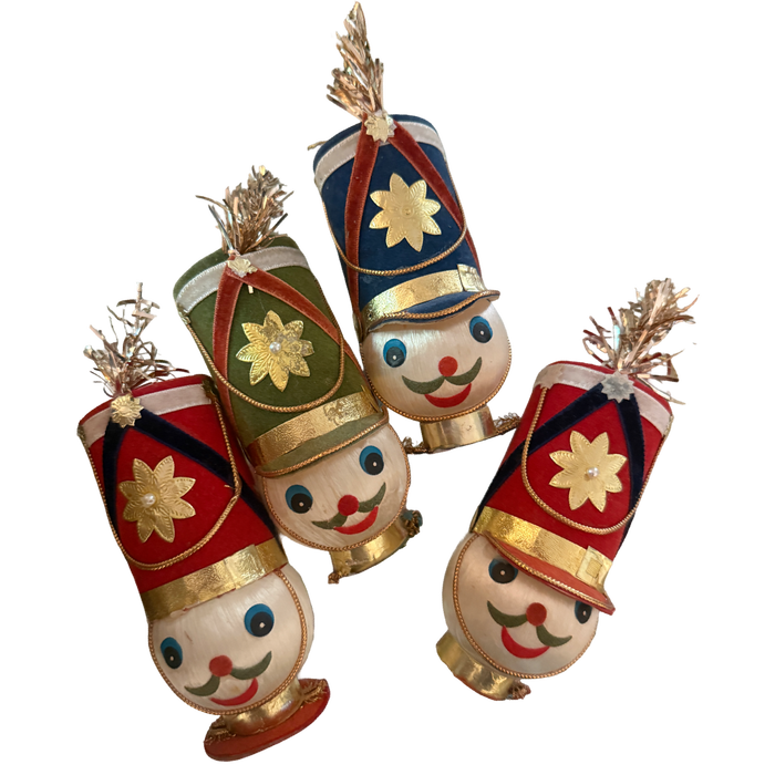 Four vintage toy soldier ornaments with smiling faces, tall red, blue, and green flocked hats, gold floral accents, and metallic tinsel plumes.