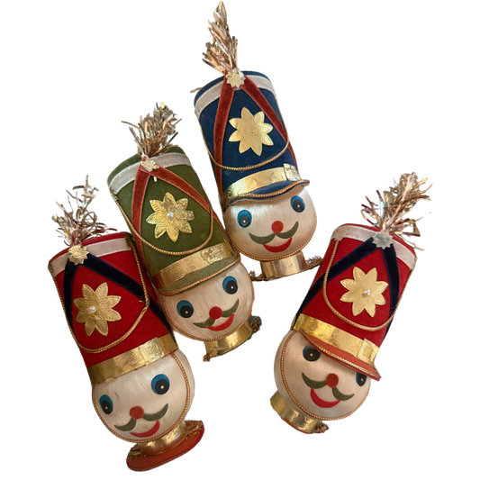 Four vintage toy soldier ornaments with smiling faces, tall red, blue, and green flocked hats, gold floral accents, and metallic tinsel plumes.