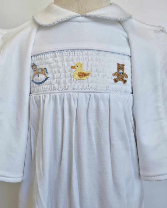 A white baby smocked dress with short sleeves, a Peter Pan collar, and a smocked chest panel featuring embroidered toys including a rocking horse, a yellow duck, and a teddy bear.