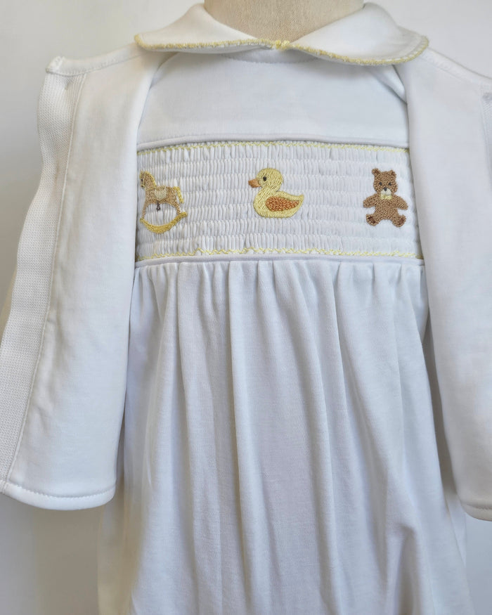 White baby smocked dress with a matching white cardigan, featuring a smocked chest panel with embroidered yellow duck, brown teddy bear, and gold rocking horse motifs.